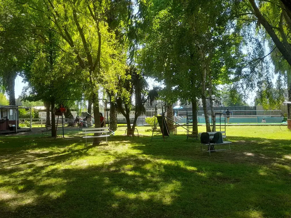 Prado arbolado con juegos y la piscina al fondo